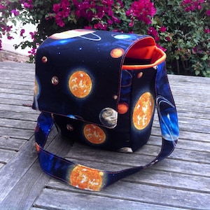 Pu&ograve; includere: Una borsa a tracolla in tessuto con una stampa spaziale blu scuro raffigurante vari pianeti, stelle e soli. La borsa &egrave; dotata di una patta, una lunga tracolla e una fodera interna arancione brillante. Il tessuto esterno presenta un motivo celeste.