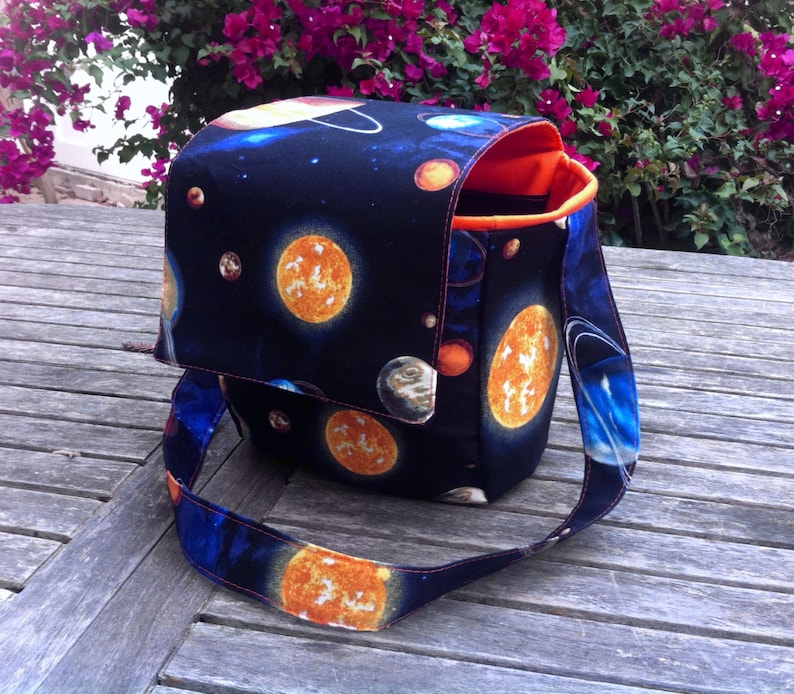 Pu&ograve; includere: Una borsa a tracolla in tessuto con una stampa spaziale blu scuro raffigurante vari pianeti, stelle e soli. La borsa &egrave; dotata di una patta, una lunga tracolla e una fodera interna arancione brillante. Il tessuto esterno presenta un motivo celeste.
