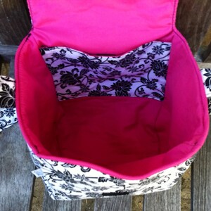 May include: A fabric bag with a black and white floral pattern on the exterior and a solid hot pink interior lining. The bag features an open top, a flap closure, and an internal pocket with the same black and white floral print design.