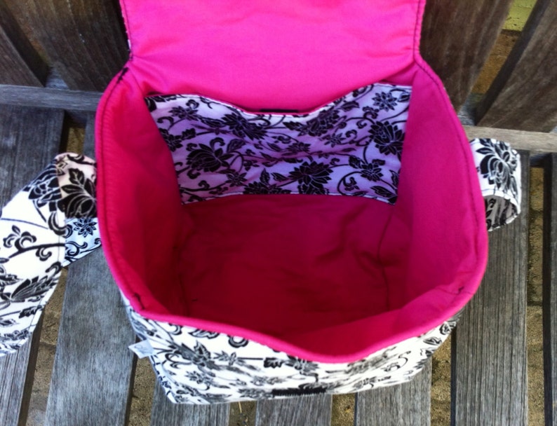 May include: A fabric bag with a black and white floral pattern on the exterior and a solid hot pink interior lining. The bag features an open top, a flap closure, and an internal pocket with the same black and white floral print design.
