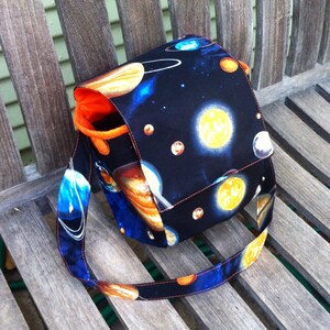 Pu&ograve; includere: Una borsa a tracolla in tessuto con un rivestimento esterno nero caratterizzato da una stampa spaziale colorata di pianeti, stelle e corpi celesti. La borsa presenta una tracolla larga e una fodera interna arancione brillante.