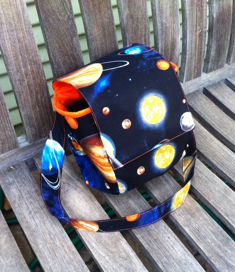 Pu&ograve; includere: Una borsa a tracolla in tessuto con un rivestimento esterno nero caratterizzato da una stampa spaziale colorata di pianeti, stelle e corpi celesti. La borsa presenta una tracolla larga e una fodera interna arancione brillante.
