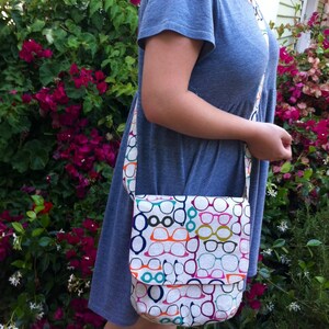 May include: A crossbody bag featuring a repeating pattern of eyeglasses in various colors including orange, teal, magenta, and dark blue. The bag has a rectangular flap closure and a matching long shoulder strap, constructed from a textured fabric.