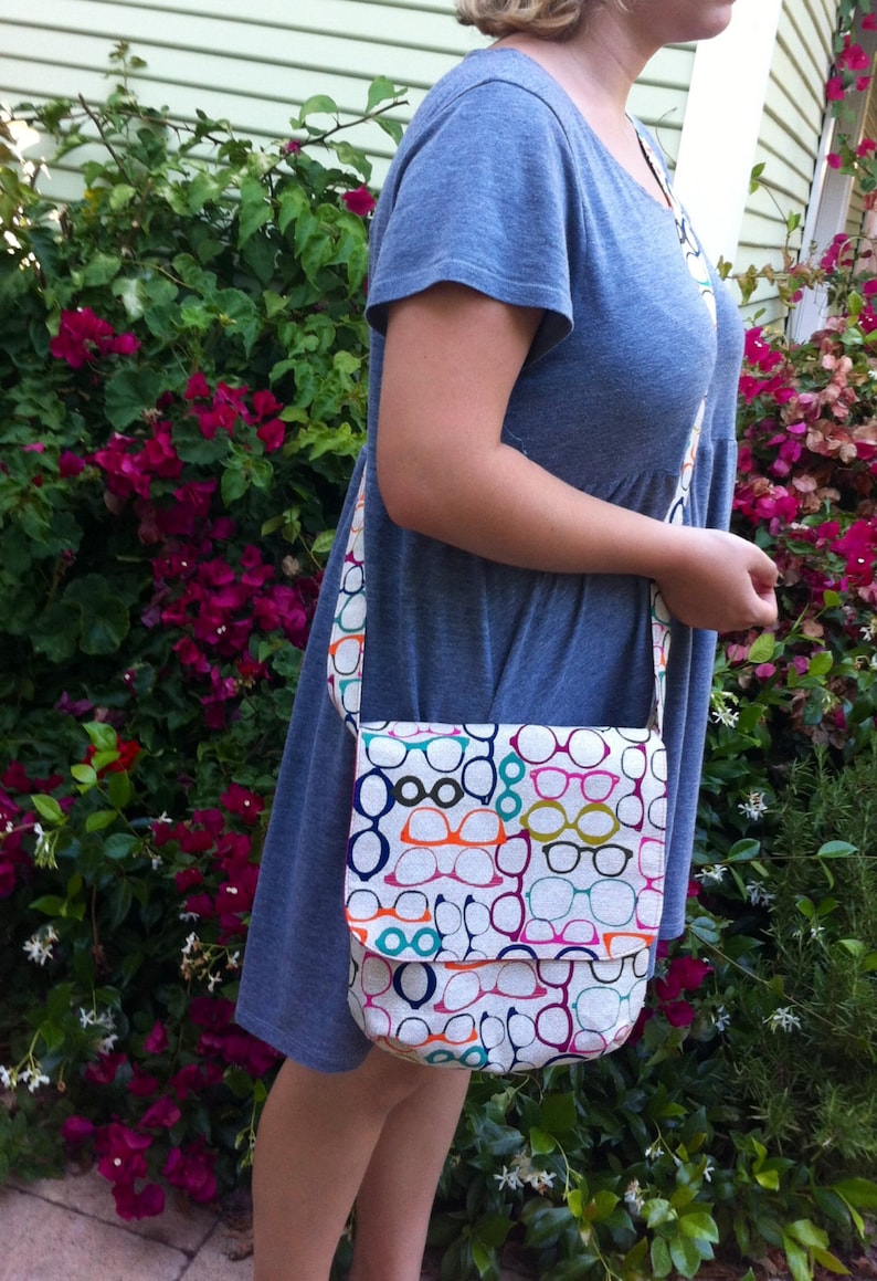 May include: A crossbody bag featuring a repeating pattern of eyeglasses in various colors including orange, teal, magenta, and dark blue. The bag has a rectangular flap closure and a matching long shoulder strap, constructed from a textured fabric.
