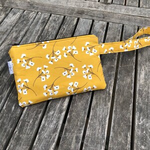 Puede incluir: Bolsa de color amarillo mostaza con un estampado floral blanco y una correa de mu&ntilde;eca a juego. La bolsa tiene una cremallera negra y una peque&ntilde;a etiqueta blanca. La tela es una mezcla de algod&oacute;n. Para objetos peque&ntilde;os.