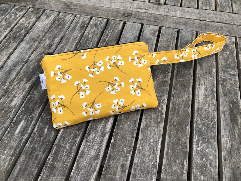 Puede incluir: Bolsa de color amarillo mostaza con un estampado floral blanco y una correa de mu&ntilde;eca a juego. La bolsa tiene una cremallera negra y una peque&ntilde;a etiqueta blanca. La tela es una mezcla de algod&oacute;n. Para objetos peque&ntilde;os.