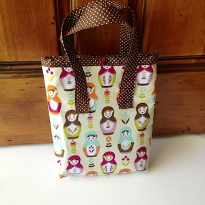 May include: A rectangular fabric tote bag featuring a repeating pattern of colourful nesting dolls and small floral motifs on a cream background. The bag has brown handles with white polka dots and a single small red button at the top centre.