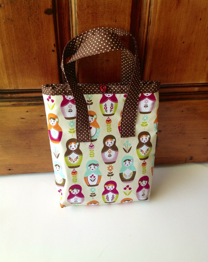 May include: A rectangular fabric tote bag featuring a repeating pattern of colourful nesting dolls and small floral motifs on a cream background. The bag has brown handles with white polka dots and a single small red button at the top centre.