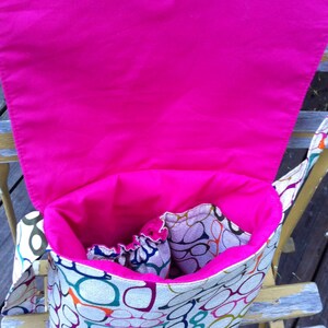 May include: A fabric messenger bag featuring a colorful pattern of eyeglasses in various shades of blue, green, orange, and purple. The bag has a bright hot pink interior lining, an open main compartment, and a flap closure with a metal snap button.