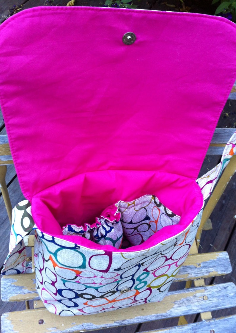 May include: A fabric messenger bag featuring a colorful pattern of eyeglasses in various shades of blue, green, orange, and purple. The bag has a bright hot pink interior lining, an open main compartment, and a flap closure with a metal snap button.
