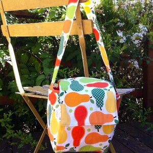 May include: A colorful shoulder bag with a pear pattern in shades of orange, yellow, and green on a white background. The bag has a flap closure and a long strap. The bag is hanging on a yellow chair outdoors.