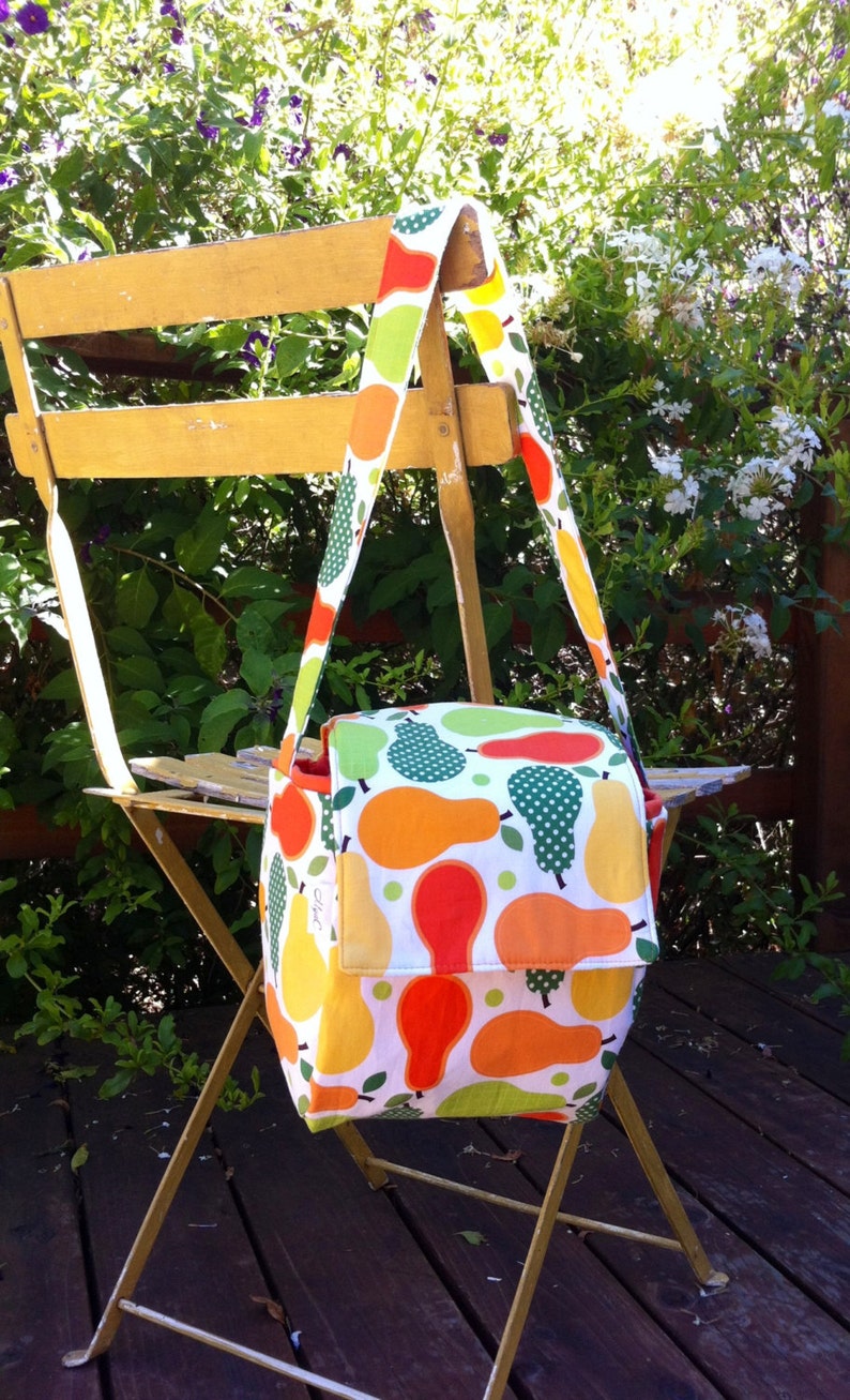 May include: A colorful shoulder bag with a pear pattern in shades of orange, yellow, and green on a white background. The bag has a flap closure and a long strap. The bag is hanging on a yellow chair outdoors.