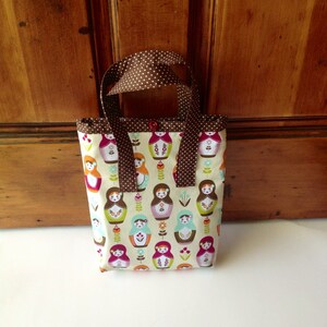 May include: A small fabric tote bag featuring a repeating pattern of colourful matryoshka dolls and small flowers on a cream background. The bag has brown handles with white polka dots and a single red button closure at the top centre opening.