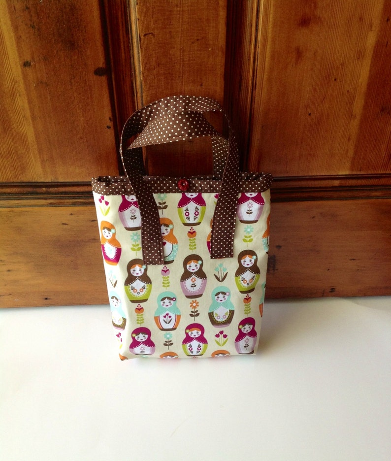 May include: A small fabric tote bag featuring a repeating pattern of colourful matryoshka dolls and small flowers on a cream background. The bag has brown handles with white polka dots and a single red button closure at the top centre opening.