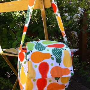May include: A colorful shoulder bag with a white background and a pattern of orange, yellow, and green pears. The bag has a flap closure and a long strap. The bag is made of fabric.