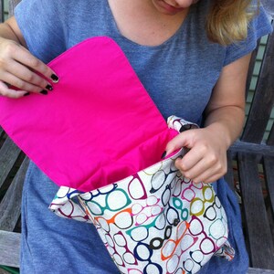 May include: A messenger bag featuring a repeating pattern of eyeglasses in various colors including orange, blue, green, and purple on a light background. The bag has a vibrant hot pink interior lining and a flap closure with a metal snap button.