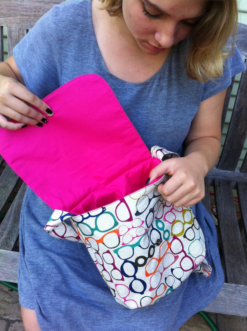 May include: A messenger bag featuring a repeating pattern of eyeglasses in various colors including orange, blue, green, and purple on a light background. The bag has a vibrant hot pink interior lining and a flap closure with a metal snap button.
