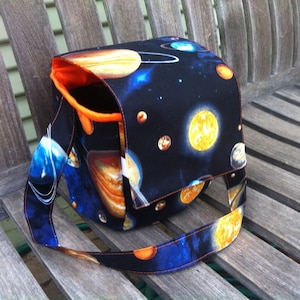 Pu&ograve; includere: Una borsa a tracolla in tessuto con sfondo nero e una stampa spaziale colorata di pianeti, stelle e corpi celesti. La borsa presenta una patta di chiusura, una lunga tracolla e una fodera interna arancione brillante visibile all'apertura.