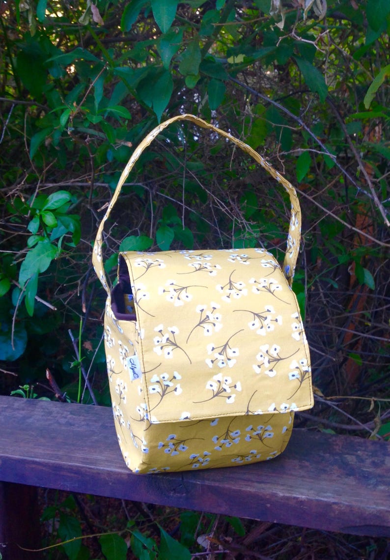 May include: A yellow fabric bag with a flap closure and a shoulder strap. The bag is decorated with a repeating pattern of white floral designs. The bag is sitting on a wooden surface, with green foliage in the background.