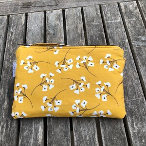 Puede incluir: Una bolsa rectangular con cremallera, con un fondo amarillo mostaza y un patr&oacute;n repetido de ramitas florales blancas. La bolsa tiene una peque&ntilde;a etiqueta y una cremallera negra.