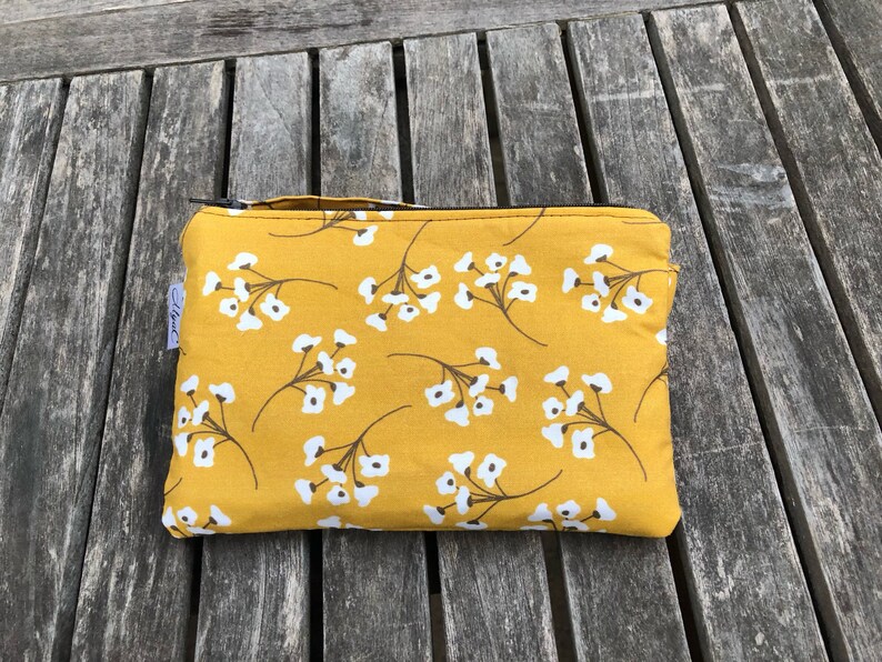 Puede incluir: Una bolsa rectangular con cremallera, con un fondo amarillo mostaza y un patr&oacute;n repetido de ramitas florales blancas. La bolsa tiene una peque&ntilde;a etiqueta y una cremallera negra.