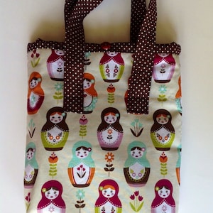 May include: A rectangular fabric tote bag featuring a repeating pattern of colourful nesting dolls and small flowers on a cream background. The bag has two brown handles with a white polka dot pattern and a single brown button closure at the top.