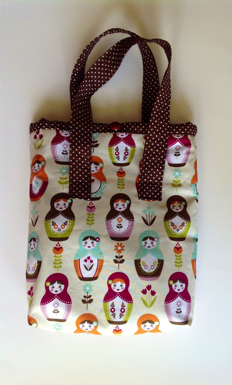 May include: A rectangular fabric tote bag featuring a repeating pattern of colourful nesting dolls and small flowers on a cream background. The bag has two brown handles with a white polka dot pattern and a single brown button closure at the top.