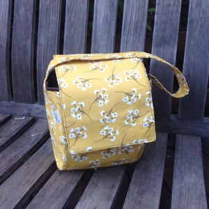 May include: A mustard yellow fabric bag with a white floral pattern. The bag has a flap closure and a handle. The bag is sitting on a wooden bench. The bag has a small tag on the side.