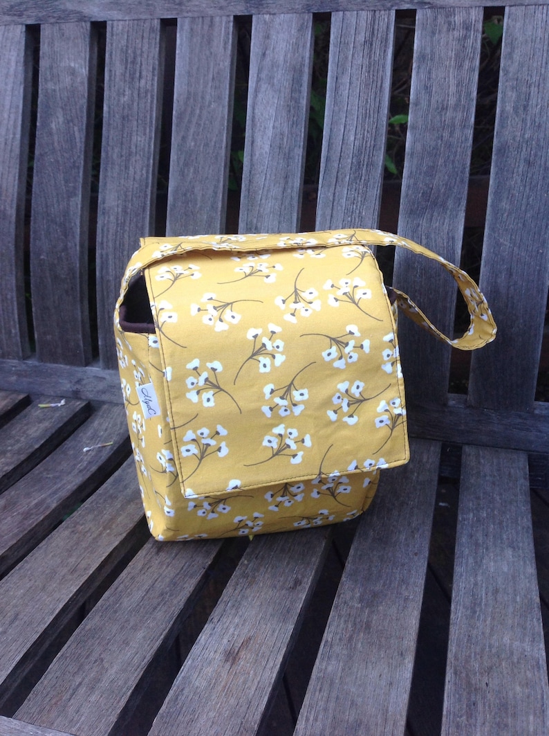 May include: A mustard yellow fabric bag with a white floral pattern. The bag has a flap closure and a handle. The bag is sitting on a wooden bench. The bag has a small tag on the side.