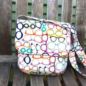 May include: A crossbody bag with a white base featuring a repeating pattern of eyeglasses in various colors including orange, pink, blue, green, and black. The bag has a fold-over flap closure and an attached shoulder strap with the same print.