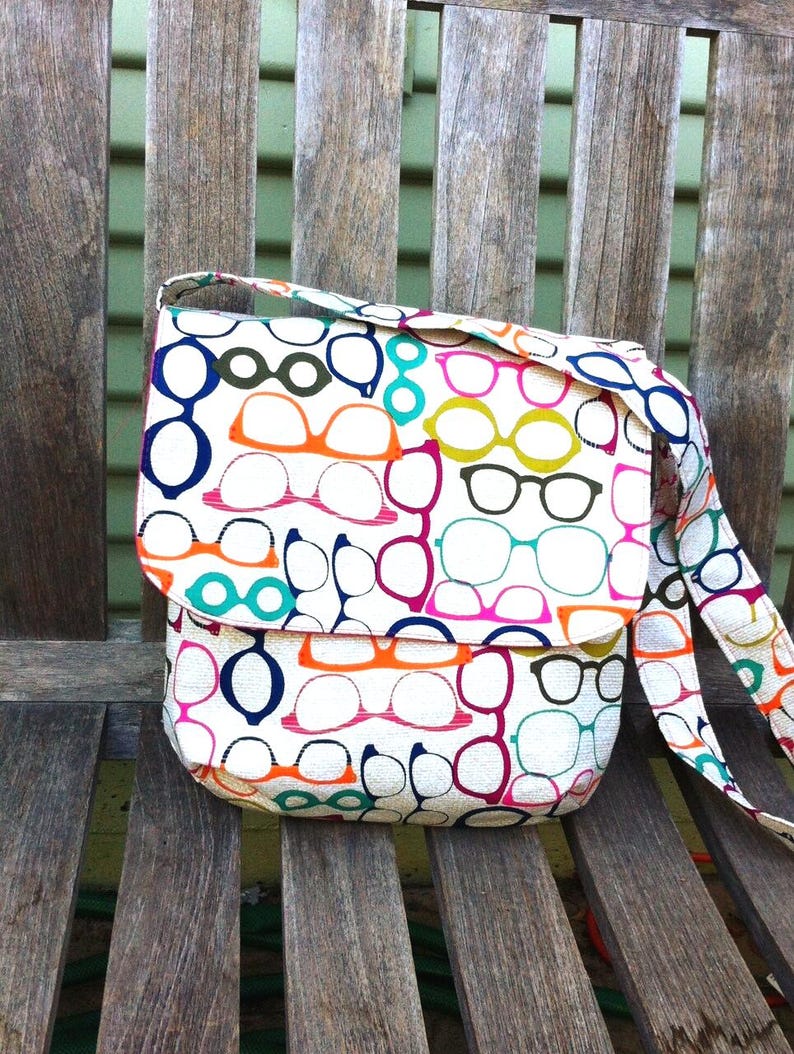May include: A crossbody bag with a white base featuring a repeating pattern of eyeglasses in various colors including orange, pink, blue, green, and black. The bag has a fold-over flap closure and an attached shoulder strap with the same print.