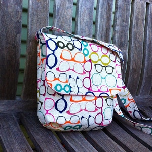May include: A rectangular shoulder bag made of off-white fabric featuring a repeating pattern of eyeglasses in various colors including orange, dark blue, magenta, and teal. The bag has a fold-over flap closure and an attached matching shoulder strap.