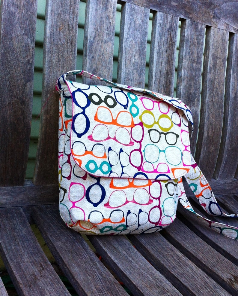 May include: A rectangular shoulder bag made of off-white fabric featuring a repeating pattern of eyeglasses in various colors including orange, dark blue, magenta, and teal. The bag has a fold-over flap closure and an attached matching shoulder strap.