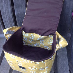 May include: Open view of a yellow floral tote bag with a brown interior. The bag has a flap closure and two handles. The interior features a brown lining and a pocket with the same floral pattern as the exterior. The bag is sitting on a wooden bench.