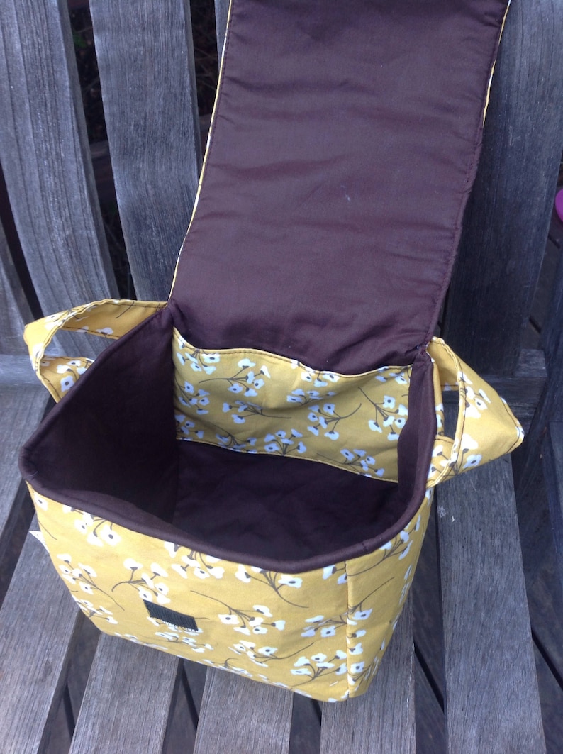May include: Open view of a yellow floral tote bag with a brown interior. The bag has a flap closure and two handles. The interior features a brown lining and a pocket with the same floral pattern as the exterior. The bag is sitting on a wooden bench.