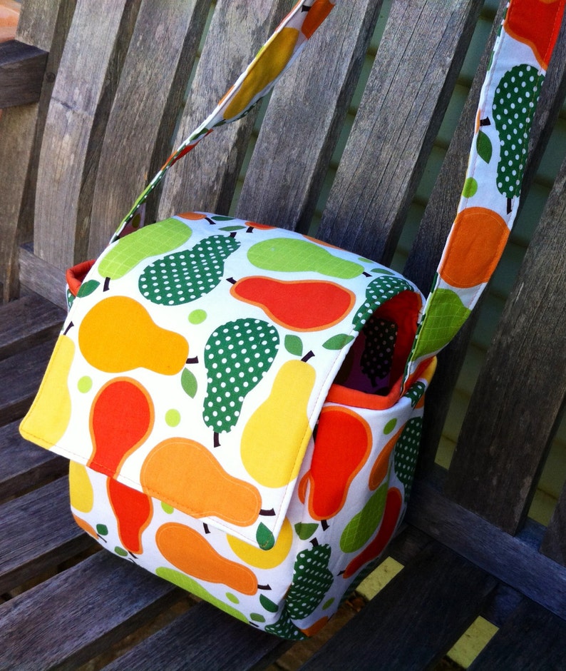 May include: A colorful crossbody bag with a flap closure, featuring a white background with a pattern of pears in shades of orange, yellow, and green. The bag has a matching strap and a red interior.