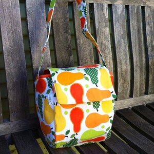 May include: A white fabric shoulder bag with a flap closure, adorned with a vibrant pattern of yellow, orange, and green pears. The bag has a matching strap and is set against a wooden bench.