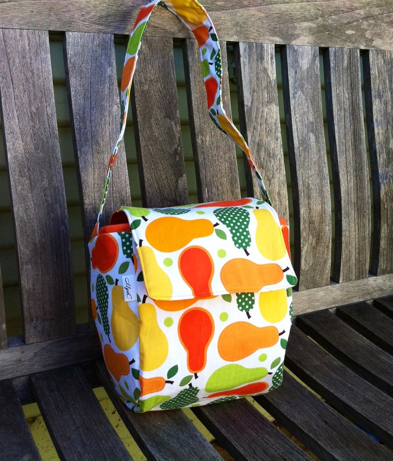 May include: A white fabric shoulder bag with a flap closure, adorned with a vibrant pattern of yellow, orange, and green pears. The bag has a matching strap and is set against a wooden bench.