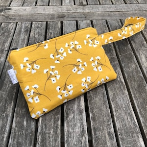 Puede incluir: Peque&ntilde;o neceser rectangular con asa, con un estampado floral amarillo mostaza con flores blancas y tallos marrones. El neceser tiene una cremallera negra y una etiqueta blanca con texto.