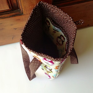May include: A fabric tote bag featuring a cream-coloured exterior with a colourful pattern of Russian nesting dolls. The interior is lined with brown fabric featuring white polka dots, and the bag includes matching brown polka-dot handles.