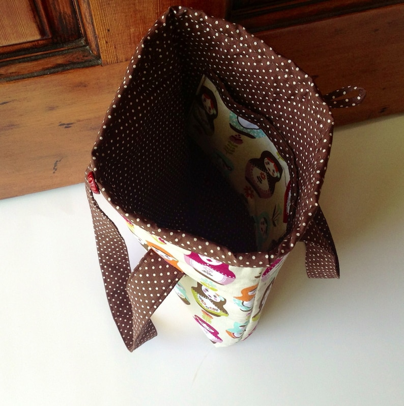 May include: A fabric tote bag featuring a cream-coloured exterior with a colourful pattern of Russian nesting dolls. The interior is lined with brown fabric featuring white polka dots, and the bag includes matching brown polka-dot handles.