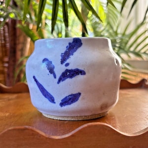 May include: A small, rounded ceramic pot with a light off-white, matte finish. The exterior is decorated with a hand-painted, abstract leaf pattern in a deep, vibrant blue color. The base of the pot is unglazed, showing a natural tan clay texture.