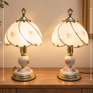 May include: A pair of table lamps with white, petal-shaped glass shades featuring floral patterns. Each lamp has a white ceramic base with matching floral designs, a gold-tone metal finish, and a height of 18 inches as indicated in the image.
