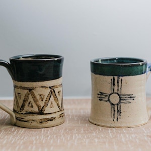 May include: Two ceramic mugs with a cream-colored base and dark blue-green rims. The left mug features a repeating brown triangular pattern, while the right mug displays a dark brown sun symbol with four radiating lines in a cross formation.