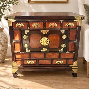 May include: A dark wood cabinet featuring a central double-door compartment with a circular brass latch, flanked by multiple drawers and panels. The piece is accented with decorative brass corner plates and hardware throughout its frame.