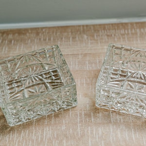 May include: Two square clear glass dishes featuring a pressed geometric pattern with starburst and grid motifs. The dishes have thick walls and a faceted exterior design that catches the light, creating a reflective and textured appearance.