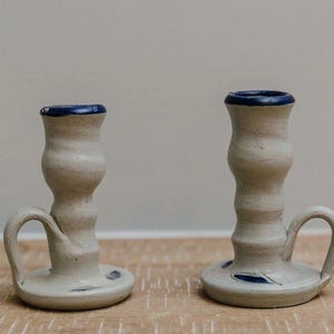 May include: Two handmade ceramic candle holders in a light beige finish with a dark blue rim. Each piece features a rounded base, a central column with a textured, undulating surface, and a small curved handle attached to the side of the base.
