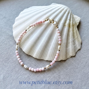 May include: A delicate bracelet featuring small, round, light pink beads interspersed with white freshwater pearls and tiny gold-toned metal spacers. The bracelet is secured with a gold-toned circular clasp and is displayed against a white seashell.