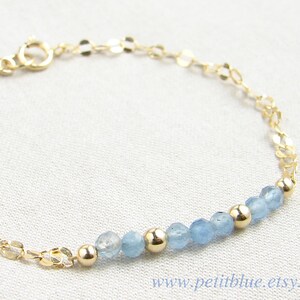 May include: A delicate gold chain bracelet with a row of light blue gemstone beads. The bracelet has a lobster clasp closure.