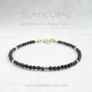 May include: A delicate bracelet made with black opal beads and gold accents. The bracelet is strung on a gold chain with a clasp closure. The text "BLACK OPAL * amplification * higher hope *" is written above the bracelet.